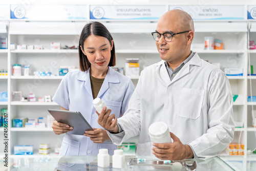 Handsome asian male pharmacist and coworker wearing lab coat and checking medicine product stock with database tablet in the pharmacy drugstore.