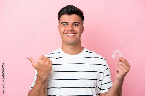 Young caucasian man holding invisaling isolated on pink background pointing to the side to present a product photo