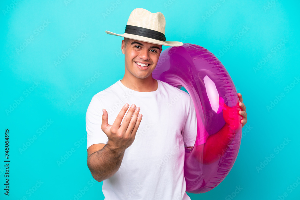 Young caucasian man holding air mattress isolated on blue background inviting to come with hand. Happy that you came Young caucasian man holding air mattress isolated on blue background inviting to come with hand. Happy that you came