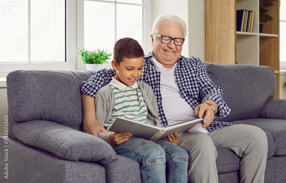 A cheerful grandfather and a child reading a book together. A happy old granddad and a little boy sitting on the sofa at home, looking at an interesting book and smiling