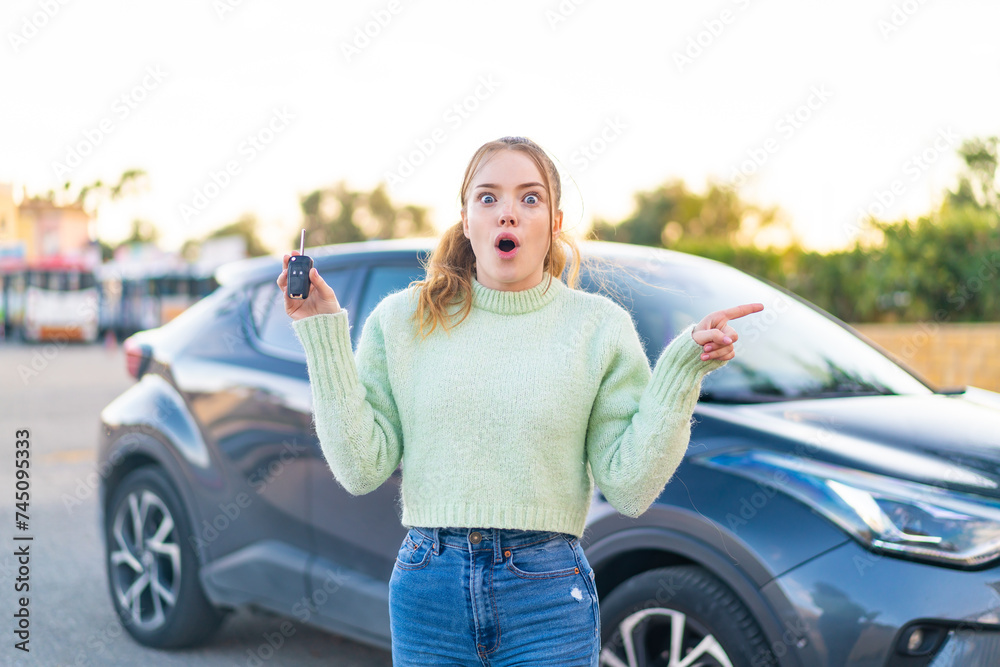 Young pretty girl holding car keys at outdoors surprised and pointing side Young pretty girl holding car keys at outdoors surprised and pointing side