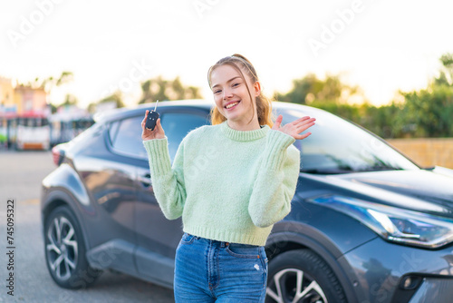 Young pretty girl holding car keys at outdoors saluting with hand with happy expression
