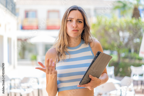 Young pretty woman holding a tablet at outdoors making doubts gesture while lifting the shoulders