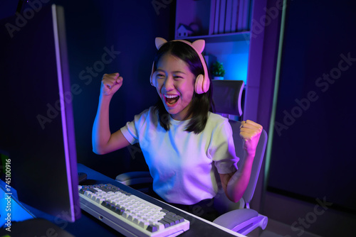 Excited and smiling Asian gamer girl in cute headset playing an online video game in her bedroom. © PaeGAG