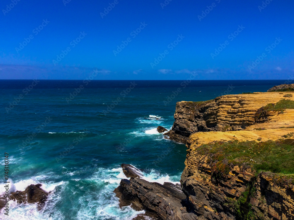 A rocky coastline with waves crashing, under a clear blue sky.