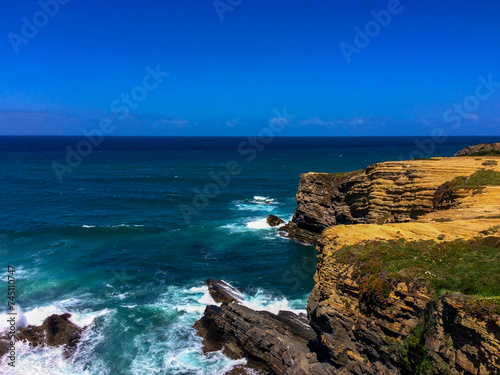 A rocky coastline with waves crashing, under a clear blue sky.