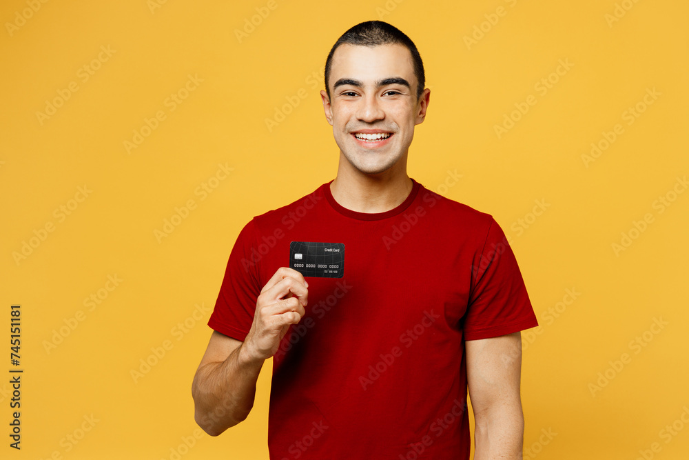 Young smiling happy fun middle eastern man he wears red t-shirt casual clothes hold in hand mock up of credit bank card isolated on plain yellow orange background studio portrait. Lifestyle concept. Young smiling happy fun middle eastern man he wears red t-shirt casual clothes hold in hand mock up of credit bank card isolated on plain yellow orange background studio portrait. Lifestyle concept.