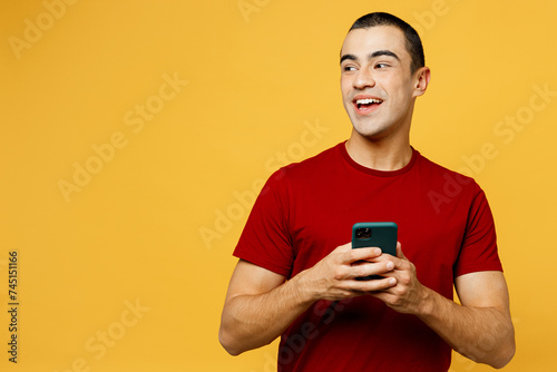 Young smiling happy middle eastern man he wear red t-shirt casual clothes hold in hand use mobile cell phone look aside isolated on plain yellow orange background studio portrait. Lifestyle concept. © ViDi Studio