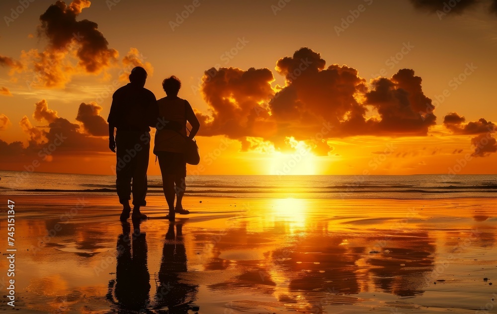 Fototapeta premium Two silhouetted figures walk on a beach at sunset the sky and clouds are reflected on the wet sand