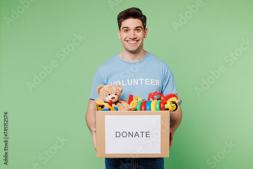 Young happy man wear blue t-shirt title volunteer hold cardboard box with donated toys look camera isolated on plain pastel green background. Voluntary free work assistance help charity grace concept.