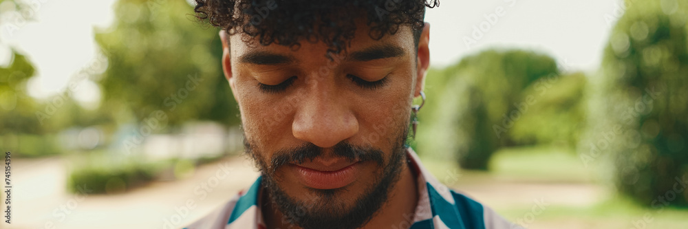 Close-up of young man with curly hair wearing striped shirt stands in park и смотрит в камеру, Panorama Close-up of young man with curly hair wearing striped shirt stands in park и смотрит в камеру, Panorama