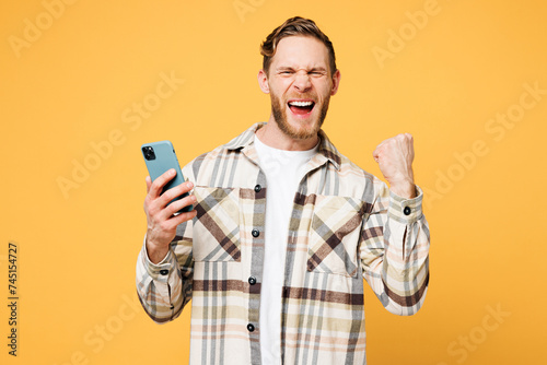 Young smiling happy Caucasian man he wear brown shirt casual clothes hold in hand use mobile cell phone chatting online do winner gesture isolated on plain yellow orange background. Lifestyle concept. photo