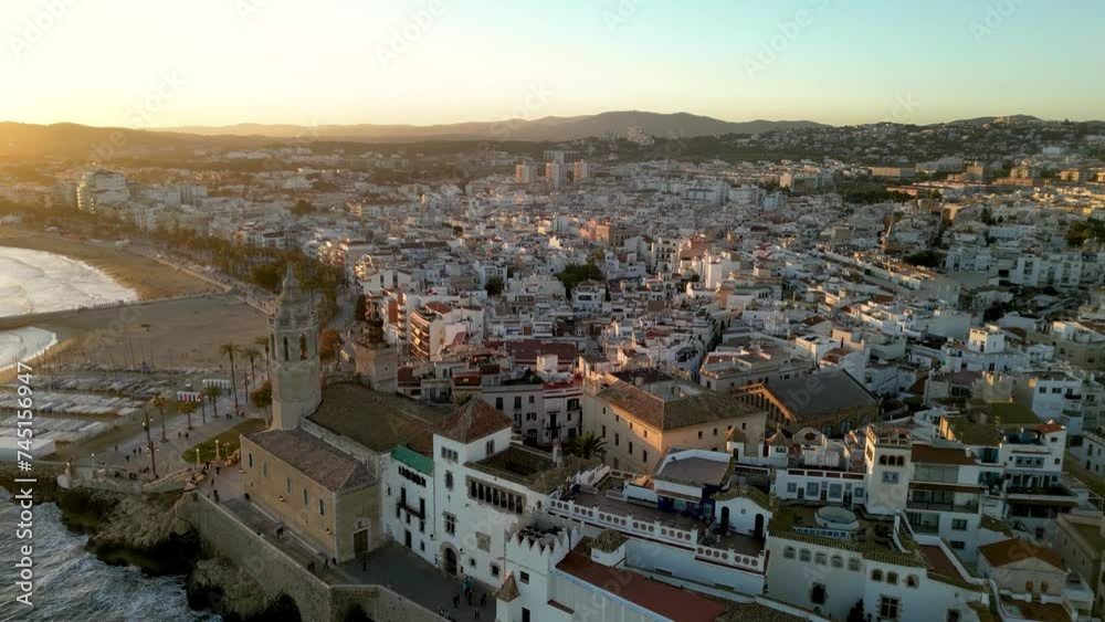 Beautiful orange sunset over Sitges city. Cinematic aerial perspective. Drone going backwards over the Church of Sant Bartolomeu i Santa Tecla. Golden hour colours. Sun reflecting on the buildings. Beautiful orange sunset over Sitges city. Cinematic aerial perspective. Drone going backwards over the Church of Sant Bartolomeu i Santa Tecla. Golden hour colours. Sun reflecting on the buildings.