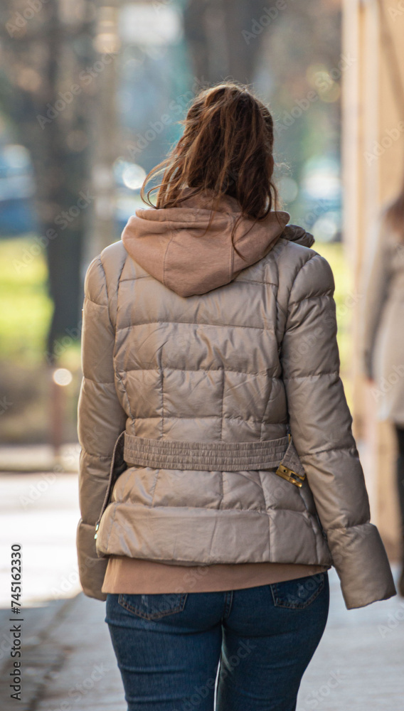 Rear view of trendy young woman in brown jacket and jeans walking on city street in spring. Female fashion concept. Back view. Trendy casual outfit. Street style