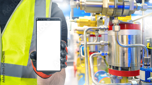 Mobile phone in hands of industrialist. Factory worker holds smartphone. Phone with blank screen. Cellphone near industrial equipment. Person in reflective vest recommends apps. Industrial mock up