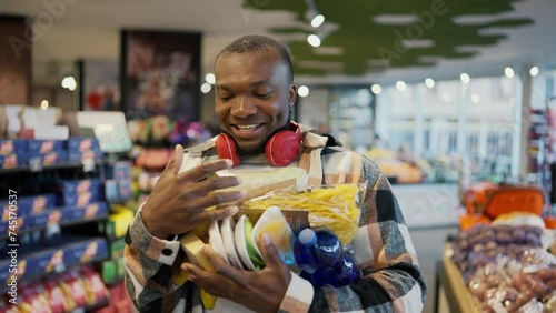 A happy and smiling man with Black skin in a checkered shirt and red wireless headphones carries a bunch of groceries on his chest holding with both hands during his walk and shopping in a modern