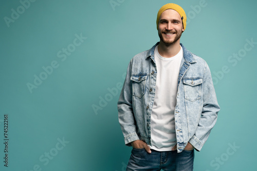 Portrait of stylish brutal young bearded European man wearing yellow hat and jeans jacket, posing at blue blank wall with copy space for your text or promotional content