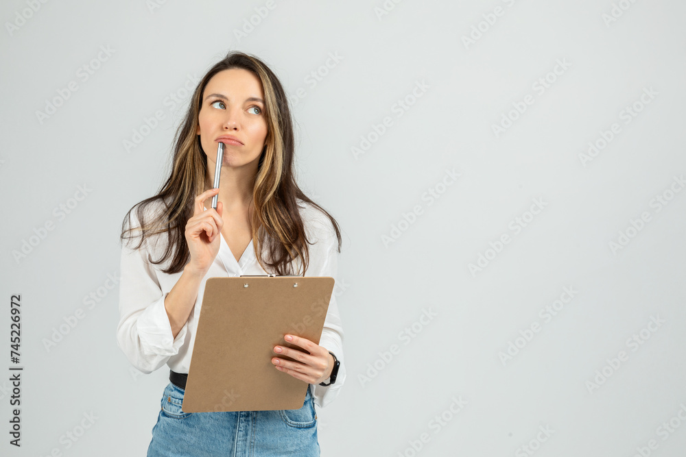 Pensive young woman holding a clipboard and a pen to her chin, looking up as if contemplating Pensive young woman holding a clipboard and a pen to her chin, looking up as if contemplating