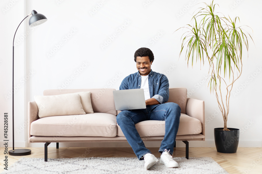 Freelance Concept. Black Man Using Laptop While Sitting On Couch At Home