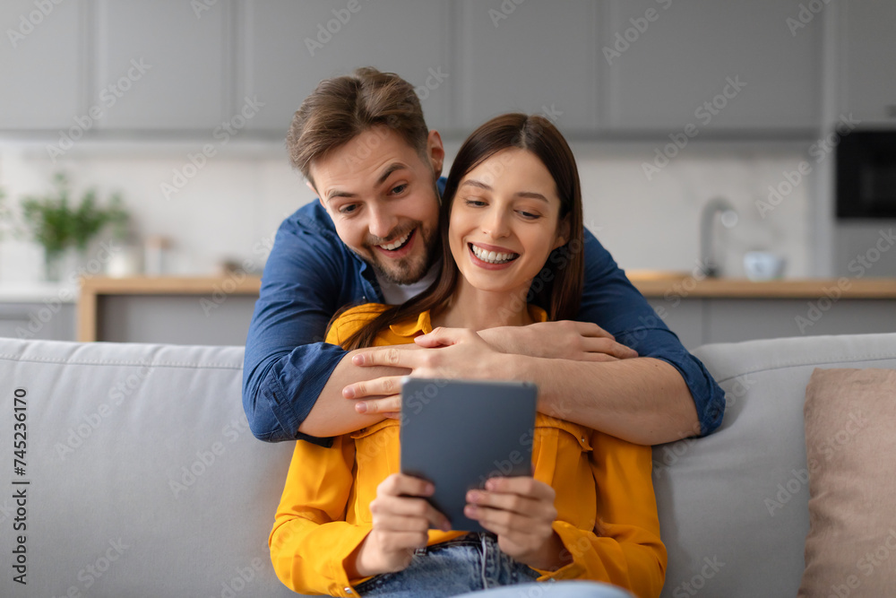 Joyful spouses with digital tablet, man hugging wife from behind Joyful spouses with digital tablet, man hugging wife from behind