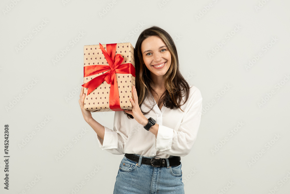 Charming woman with a beaming smile holding a beautifully wrapped gift with a red bow Charming woman with a beaming smile holding a beautifully wrapped gift with a red bow
