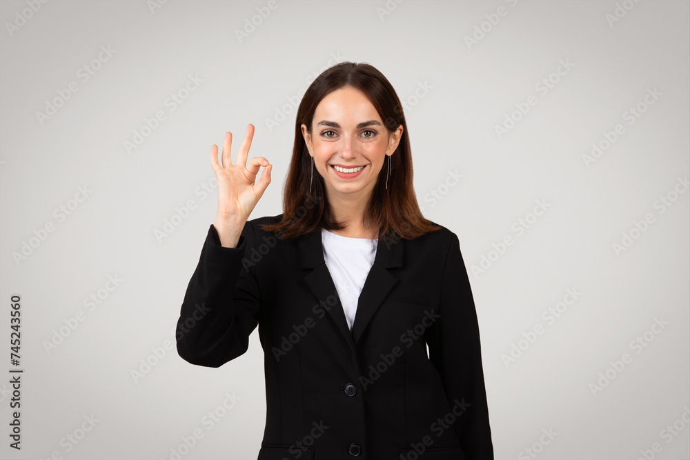 Cheerful young businesswoman in a smart black suit making a positive 'okay' hand sign Cheerful young businesswoman in a smart black suit making a positive 'okay' hand sign