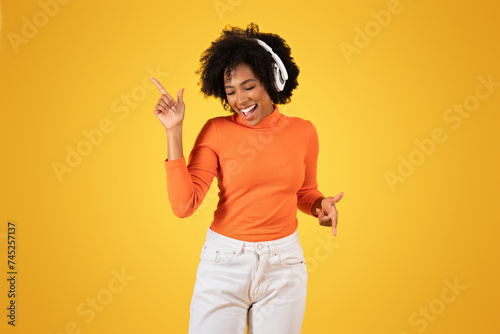 Joyful young woman with curly afro hair wearing headphones dances and points upwards