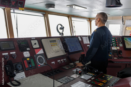 Officer on watch on the navigational bridge. Caucasian man in blue uniform sweater on the bridge of cargo ship.