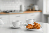 Cups of coffee and fresh croissant on white table with blurred kitchen