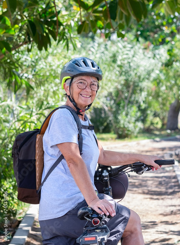 Active smiling senior woman cycling with electro bike in a green park wearing helmet looking back. Mature attractive woman enjoying retirement and healthy lifestyle.