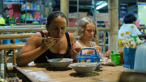 Young woman with little daughter eating in Traditional Thai cafe, tasting Guay teow noodle beef soup photo