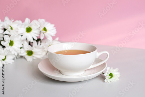 A white cup with tea and a saucer stands on a table with white chrysanthemum flowers