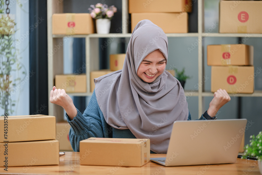 Active asian woman in blue muslim suit sitting and working with computer and online package box delivery. Startup small business freelance working girl showing thumb up with exciting emotion. Active asian woman in blue muslim suit sitting and working with computer and online package box delivery. Startup small business freelance working girl showing thumb up with exciting emotion.
