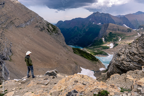 Matured Caucasian man standing at the edge of a cliff watching a glacial lake in a deep canyon with snow high mountains all around, garden Wall, Grinnell Lake, Glacier National Park, Montana