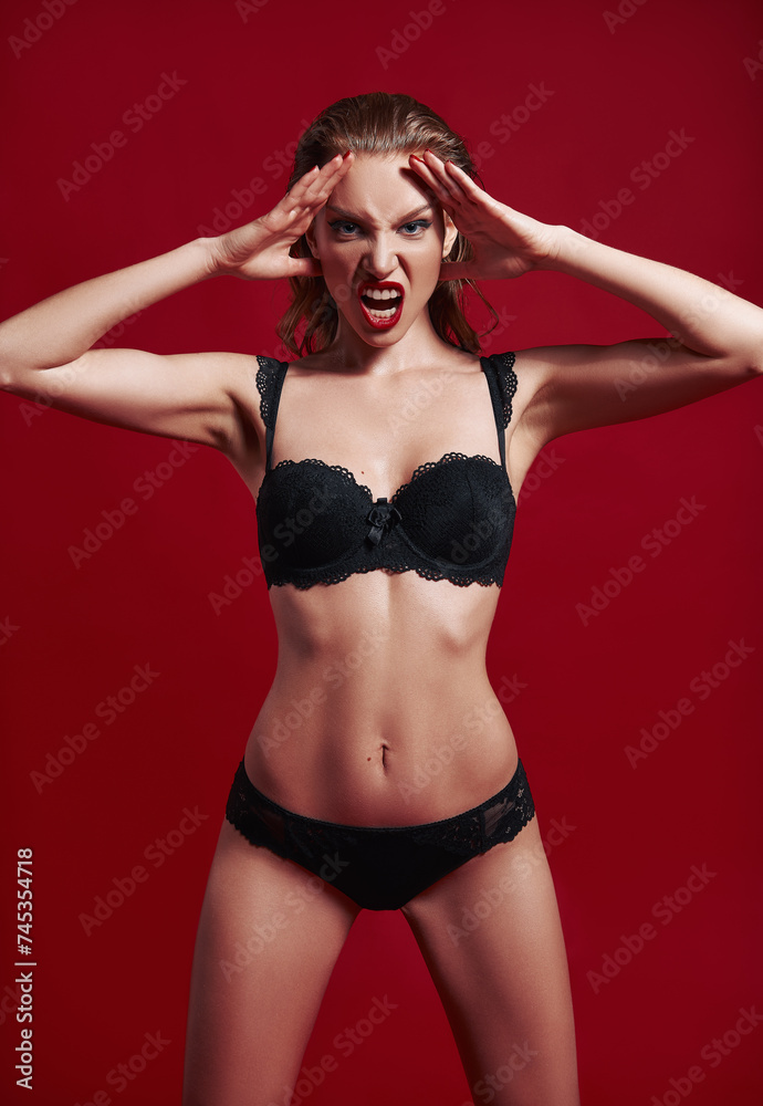 Studio fashion shot of sexy angry young woman. Portrait of screaming beautiful girl dressed in black lingerie. Seductive evil lady against red background Studio fashion shot of sexy angry young woman. Portrait of screaming beautiful girl dressed in black lingerie. Seductive evil lady against red background