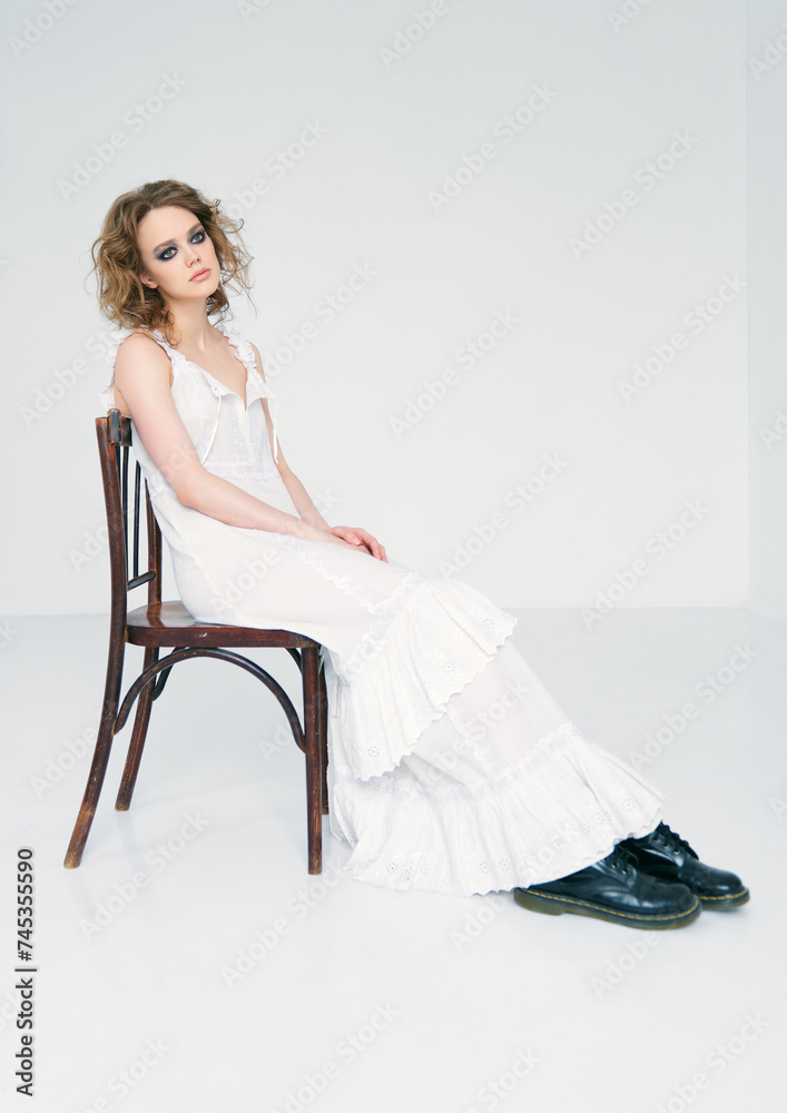 Fototapeta premium Portrait of pretty strange sad girl. Beautiful weird young woman in white dress sitting on stool in empty room