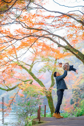Happy Asian father rising up smiling baby girl with colorful trees in autumn season in background 