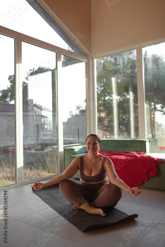 A beautiful plus size girl does yoga at home. The woman is wearing a tracksuit, leggins and a rop and posing against the background os a panoramiv windows and a sofa photo