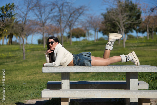 Young, beautiful, brunette woman in a white sweater, denim miniskirt and sunglasses, funny, happy and relaxed, lying on a park bench on a sunny day. Beauty, fashion, trend concept.