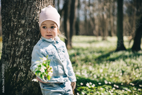 Little girl in jeans jacket standing near the tree in the forest covered with anemones 