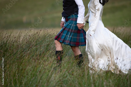 A newly married Scottish couple strolls through a grassy field in Glencoe, the Scottish highlands. He dressed in a Scottish Kilt and she dressed in a white wedding dress photo