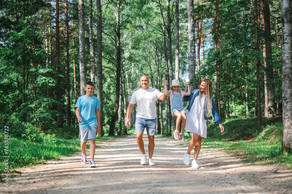 Young Family of four enjoying a walk in the forest, parents swinging the daughter, and the teenage children strolling alongside. for themes of family bonding and nature outings. Young Family of four enjoying a walk in the forest, parents swinging the daughter, and the teenage children strolling alongside. for themes of family bonding and nature outings.