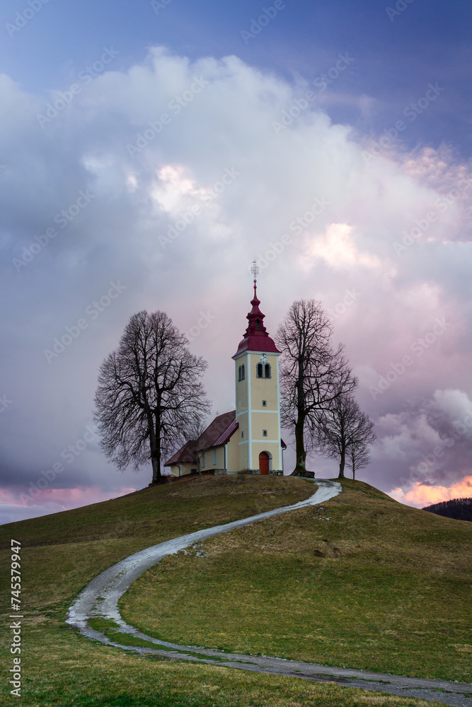 A path to the church on the hil in the clouds. A scenic church in the sunset pinkish clouds. An ancient church surrounded by leafless trees. A church of saint Thomas in Gorenji Vrsnik - Slovenia. 
