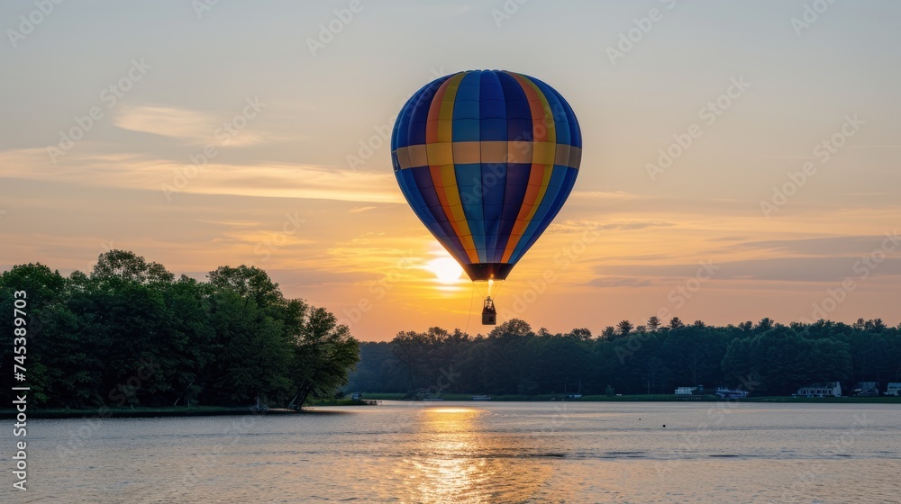 Obraz premium a hot air balloon flying over a body of water with the sun setting in the background and trees in the foreground.