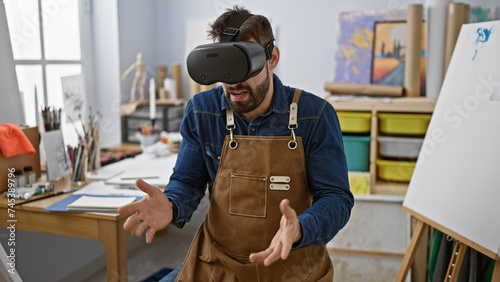 Young, bearded hispanic artist in apron manically draws on canvas in art studio using futuristic virtual reality glasses