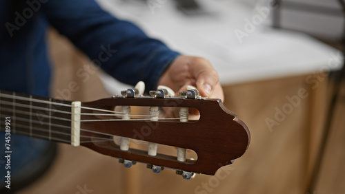 Handsome young hispanic man passionately tuning his classical guitar, setting the stage for a melodic performance in the heart of a cozy music studio