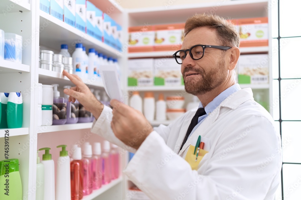 Middle age man pharmacist holding product on shelving reading prescription at pharmacy