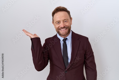 Middle age business man with beard wearing suit and tie smiling cheerful presenting and pointing with palm of hand looking at the camera.