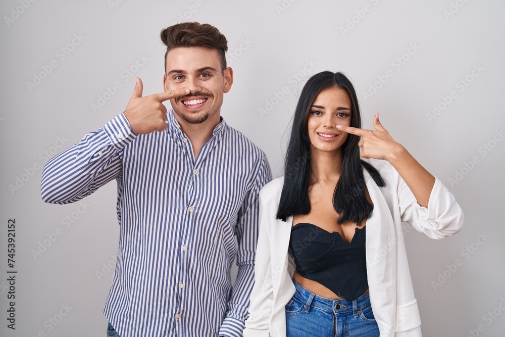 Young hispanic couple standing over white background pointing with hand finger to face and nose, smiling cheerful. beauty concept
