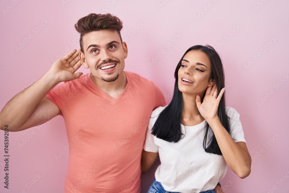 Young hispanic couple standing over pink background smiling with hand over ear listening an hearing to rumor or gossip. deafness concept. Young hispanic couple standing over pink background smiling with hand over ear listening an hearing to rumor or gossip. deafness concept.